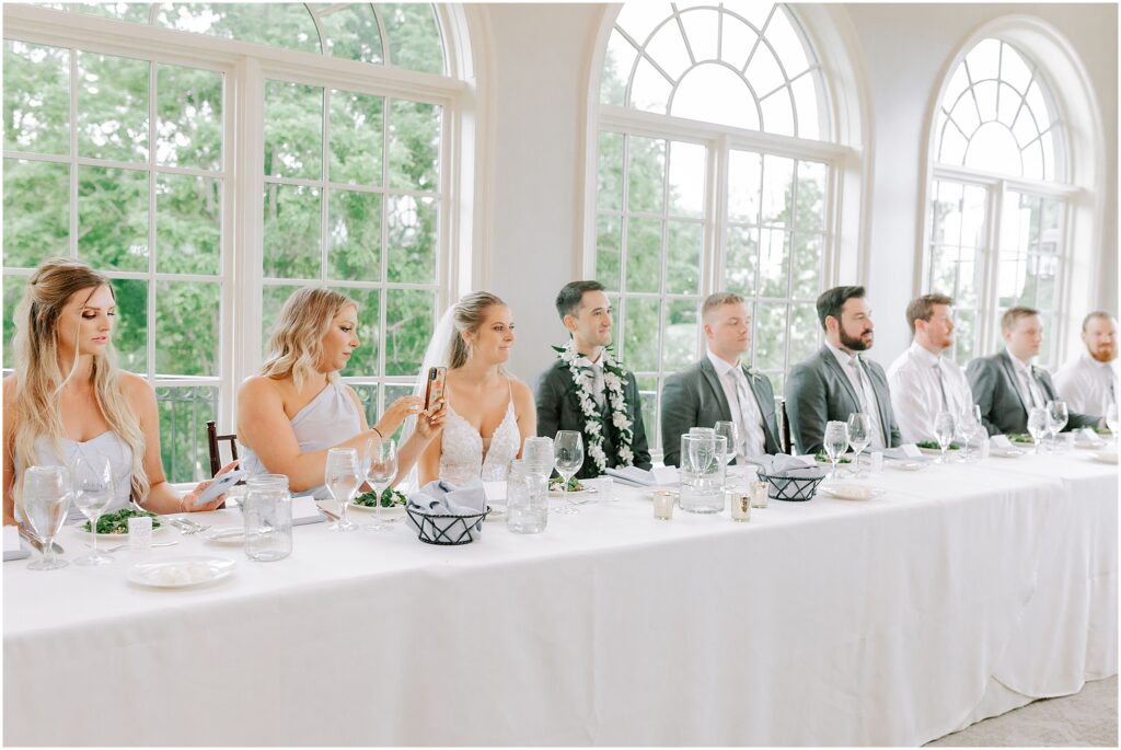 wedding party sits at long all white head table inside reception at Stonewall Golf Club
